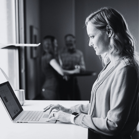 A black and white image of a lady typing on a laptop standing at a desk with two people having coffee in the background